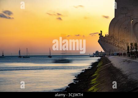 Denkmal für Entdeckungen bei Sonnenuntergang entlang des Tejo, Belém, Lissabon, Portugal mit einem Segelboot, das den Hafen vor goldenem Himmel in der Silhouette einfährt Stockfoto