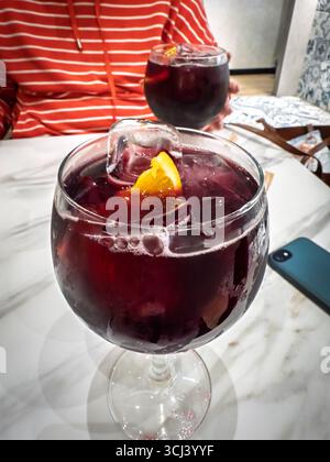 Nahaufnahme von zwei Gläsern roter Sangria mit Eiswürfeln und orangefarbenen Keilen auf einem Marmortisch in einer Bar in Valencia. Kondenswasser schimmert auf dem Glas, während ein Ba Stockfoto