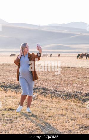 Schwangere Frau macht ein Selfie mit einem Telefon, steht auf einem großen Feld mit Pferden im Hintergrund an einem sonnigen Tag. Stockfoto