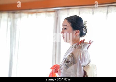 Eine Frau im Kimono steht vor einem Fenster. Sie trägt einen roten Schal und hat ihr Haar in einem Brötchen. Konzept von Eleganz und Tradition Stockfoto