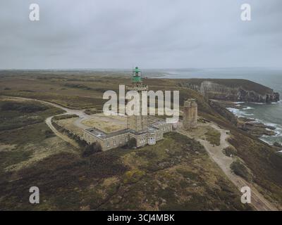 Aus der Vogelperspektive erhebt sich der Leuchtturm Phare du Cap Fréhel vor der zerklüfteten Küste, wo das smaragdgrüne Meer auf die ockerfarbenen Klippen trifft, Plévenon, Bretagne, Frankreich. Stockfoto