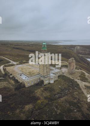 Aus der Vogelperspektive des Leuchtturms Phare du Cap Fréhel, der hoch über der gedämpften Landschaft steht und dessen grüne Spitze im Kontrast zum düsteren Himmel steht, Plévenon, Bretagne, Frankreich. Stockfoto