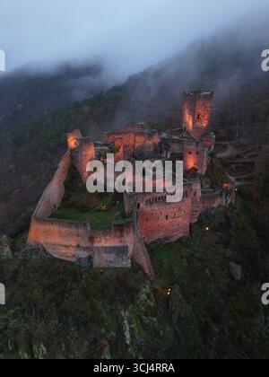 Aus der Vogelperspektive auf die Château de Saint-Ulrich, eine mittelalterliche Burgruine, die warm vor dem dunklen, nebeligen Hügel leuchtet, Ribeauvillé, Grand Est, Frankreich. Stockfoto