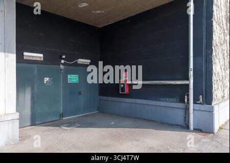 Zwei graublaue Garagentore aus Stahl stehen in der Ecke eines dunkelmaurigen Gebäudes. Über den Türen befinden sich zwei Lichter und eine Überwachungskamera. Ein roter Feuerausgang Stockfoto