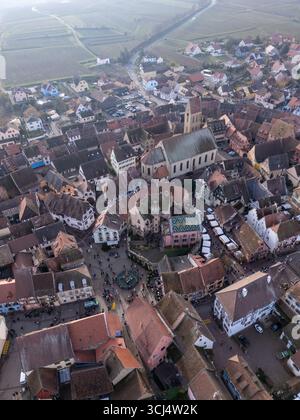 Aus der Vogelperspektive auf die Terrakotta-Dächer, die sich um die zentrale Kirche und den Marktplatz kaskadieren, eingebettet zwischen Weinbergen unter einem weichen, trüben Himmel, Eguisheim, Grand Est, Frankreich. Stockfoto