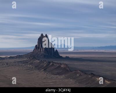 Aus der flachen, trockenen Wüstenlandschaft unter einem riesigen Himmel erhebt sich Shiprock, New Mexico, USA, dramatisch aus der Vogelperspektive von Shiprocks zerklüfteter, dunkler vulkanischer Felsformation. Stockfoto
