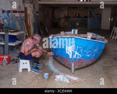 Der Mann repariert und lackiert ein kleines Boot in einer Werkstatt und zeigt traditionelle Bootswartungsarbeiten. Stockfoto