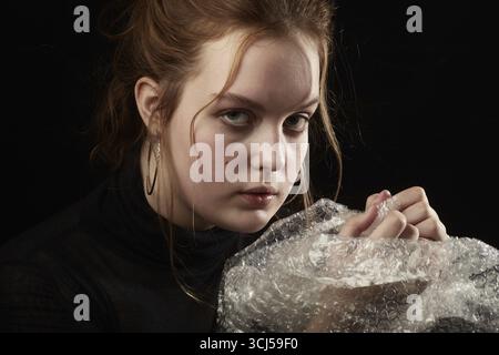 Traurige junge Frau mit Down-Syndrom, die Plastikfolie auf schwarzem Hintergrund platzt. Anti-Stress Stockfoto