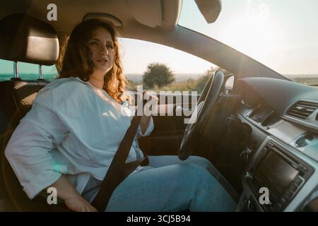 Junge Frau, die bei Sonnenuntergang im Auto den Sicherheitsgurt anlegt Stockfoto