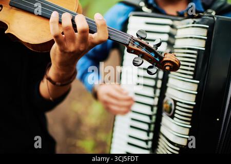 Kaukasische junge Erwachsene Frau, die Violine spielt, neben kaukasischem jungen erwachsenen Mann, der im Freien Akkordeon spielt, Hände und Instrumente im Fokus, Musiker, die in unmittelbarer Nähe auftreten Stockfoto