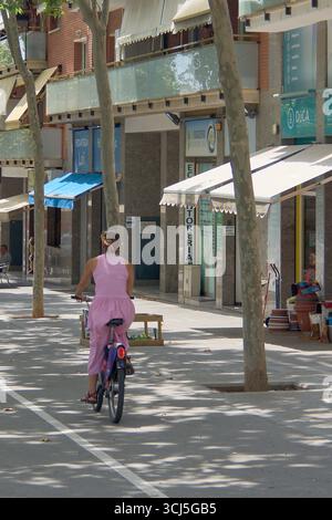 Viladecans. Barcelona - 05. September 2025: Eine Frau in elegantem rosa Kleid fährt auf einer von Bäumen gesäumten Fußgängerzone mit dem Fahrrad. Die Szene ruft hervor Stockfoto