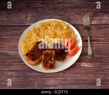 Frittierte Fleischkoteletts auf einem runden Teller mit Reifen Tomatenscheiben, Makkaroni und geriebenem Käse auf einem Holztisch. Draufsicht, flach. Stockfoto