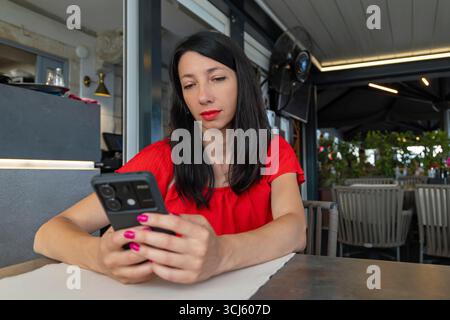 Frau in rotem Kleid mit Smartphone im Café. Eine brünette Frau in einem roten Kleid sitzt an einem modernen Café-Tisch im Freien und konzentriert sich dabei auf ihr Smartphone Stockfoto