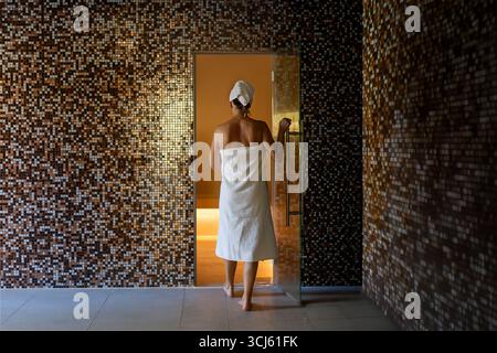 Eine Frau betritt ein luxuriöses türkisches Bad in einer warmen und entspannenden Atmosphäre. Die Wände sind mit herrlichen Mosaiken bedeckt, die das Licht reflektieren. Stockfoto