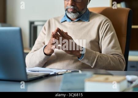 Indischer Mann mittleren Alters, der am Schreibtisch sitzt und die Hände vor dem Laptop hält, während der Fernarbeit oder beim Online-Meeting nachdenklich wirkt, Brille und Bart trägt Stockfoto