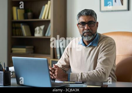 Porträt eines südasiatischen Mannes mittleren Alters mit grauen Haaren und Bart, der am Schreibtisch sitzt, eine Brille trägt, in die Kamera schaut, im Hintergrund ein Bücherregal Stockfoto