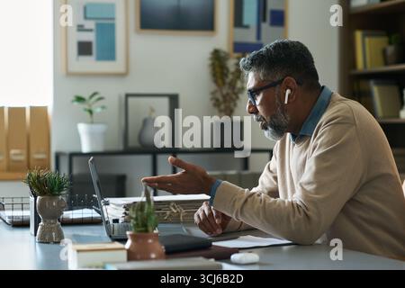 Indischer Mann mittleren Alters mit grauem Bart, der am Schreibtisch sitzt und beim Videoanruf auf dem Laptop Gesten macht, Brille und schnurlose Ohrhörer trägt, moderner Bürohintergrund sichtbar Stockfoto