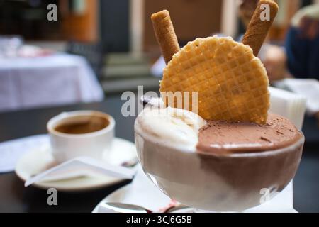 Ein Glas gefüllt mit frisch zubereitetem Eis mit Vanille- und Schokoladengeschmack mit einem runden Waffel und Rollen auf einem Tisch in einem Café, eine Tasse Kaffee im Stockfoto