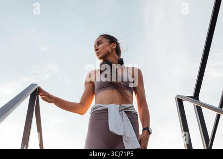 Niedrigwinkelansicht der jungen, schlanken Frau in Sportbekleidung, die auf der Treppe auf dem Dach steht und wegblickt Niedrigwinkelansicht der jungen, schlanken Frau in Sportbekleidung Stockfoto