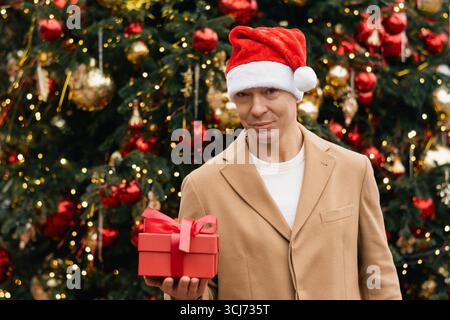Glücklicher kaukasischer Mann in beigem Mantel mit rotem Geschenk neben dem Weihnachtsbaum. Weihnachtsstimmung. Stockfoto