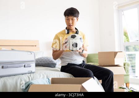 Die Kisten im Schlafzimmer auspacken, ein junger Mann, der Fußball mit Kopfhörern hält Stockfoto