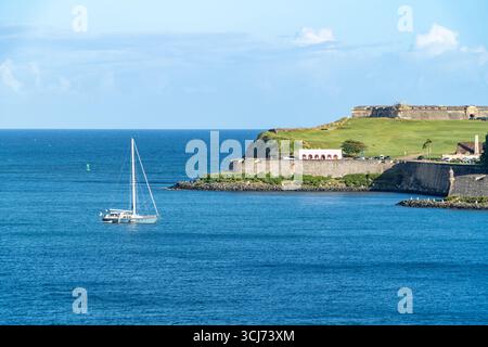 Ein einsames Segelboot fährt über ruhiges blaues Wasser in der Nähe einer Landzunge mit historischen Steinbefestigungen. Heller, sonniger Tag mit klarem Himmel an einer tropischen Küste Stockfoto