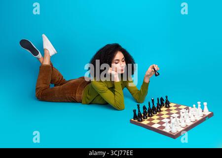 Junge Frau, die Schach spielt, während sie auf einem leuchtend blauen Hintergrund liegt, zeigt Freizeit und nachdenkliche Aktivität Stockfoto