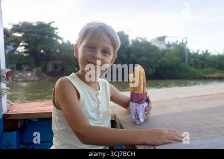 Kind, isst Sandwich auf einer Bootsfahrt in Vietnam, im Sommer Stockfoto