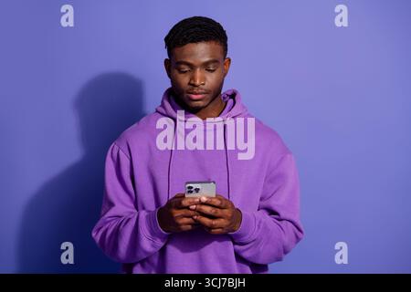 Ein junger Mann in einem violetten Hoodie benutzt ein Smartphone, steht vor einem violetten Hintergrund und zeigt stilvolle Freizeitmode Stockfoto