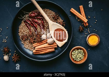 Gemischte Bio-Gewürze (Koriander, Muskatnuss, Anissterne, Cayennepfeffer, Kurkuma, Paprika, Zimtstangen) auf Keramikplatte auf schwarzem Hintergrund, oben Stockfoto