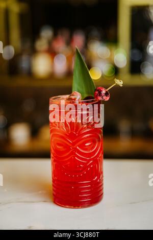 Tropischer roter Cocktail mit Kirschen und Blattgarnitur in einem Tiki-Glas an einer Bar Stockfoto