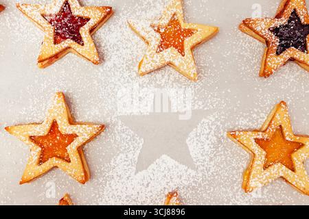 Sternförmige Kekse, gefüllt mit verschiedenen Marmeladen, sind auf einem Backblech angeordnet, das mit Puderzucker bestäubt ist. Die Kekse sorgen für eine festliche und einladende Atmosphäre Stockfoto