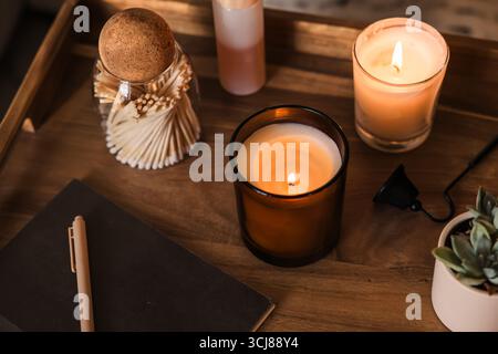 Eine warme, einladende Szene mit zündeten Kerzen, Notizblock zum Journalisieren, einem Glas Streichhölzer, einem Notizbuch mit Stift und einer kleinen Pflanze auf einem Holztisch. Stockfoto