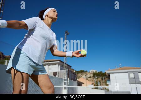 Eine entschlossene junge Frau konzentriert sich darauf, einen Tennisball auf einem Platz im Freien zu servieren. Ihre sportliche Haltung und die sonnige Kulisse betonen die aktive Aktivität Stockfoto