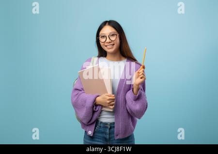 Ich kenne die Antwort. Aufgeregt asiatische Studentin in Brillen halten Notizblock und zeigen Bleistift nach oben, blauer Hintergrund Stockfoto