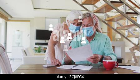 Senioren mit chirurgischen Masken, die Dokumente am Esstisch überprüfen, mit Notebook-Stift-Kaffeetasse Stockfoto