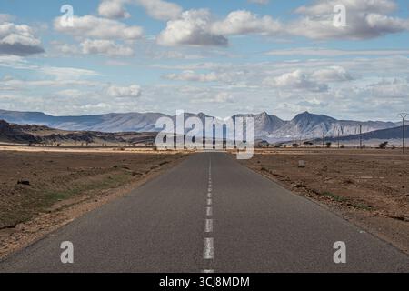 Lange Straße durch die marokkanische Wüste, die zu entfernten felsigen Bergen mit flachem kargem Land auf beiden Seiten unter hellem Himmel mit verstreuten Wolken führt Stockfoto