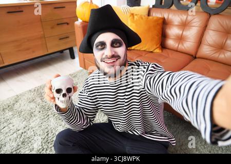 Junger glücklicher Mann, verkleidet als Pirat für Halloween mit menschlichem Schädel, der Selfie im dekorierten Wohnzimmer macht Stockfoto