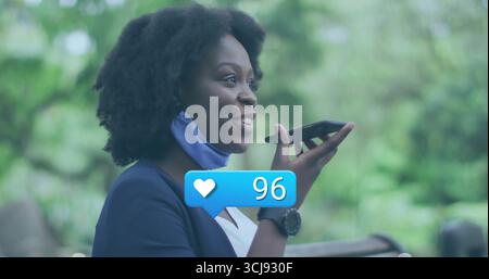Frau mit Blazer, Smartwatch, Maske am Kinn, Smartphone auf der Bank im Park, mit Like-Symbol Stockfoto