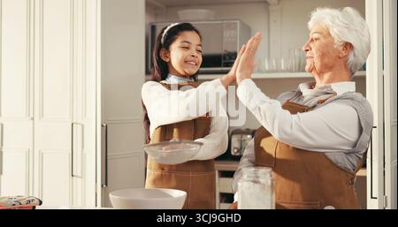 Glückliche Großmutter, Enkelkind oder Backen mit High Five in der Küche für Erfolg oder Dessertrezept zu Hause. Ältere Frau, Mädchen oder Kind mit Lächeln, Mehl oder Stockfoto