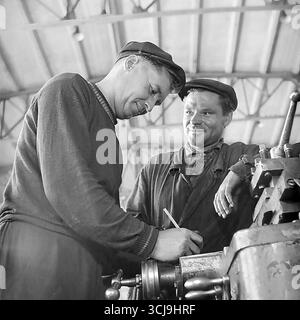 Zwei Arbeitergenossen diskutieren eine Aufgabe an einer Drehmaschine in einer Fabrikwerkstatt in Sloviansk, 1970er Jahre Das Archivfoto vermittelt eine warme Atmosphäre der Freundschaft und Mentorität in einem sowjetischen Werkkollektiv. Diese Szene friedlicher, kreativer Arbeit und menschlicher Beziehungen ist ein Symbol für das Leben im Donbass vor dem Krieg Stockfoto