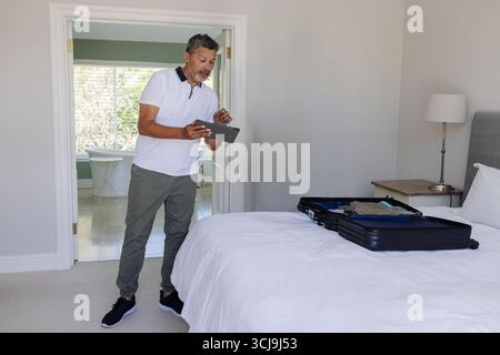 Mann, der ein digitales Tablet hält, während er neben dem Koffer auf dem Bett im Hauptschlafzimmer in der Nähe der Lampe steht Stockfoto