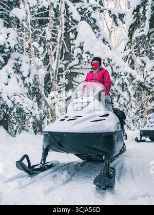 Eine junge Frau in warmer Winterkleidung navigiert mit einem Schneemobil auf einem verschneiten Pfad, der von hohen Bäumen umgeben ist. Stockfoto