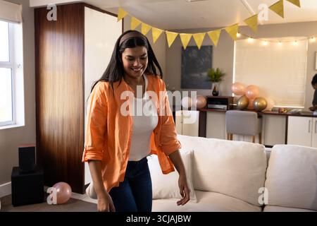 Lächelnde Frau in orangefarbenem Hemd, die festliche Heimfeier mit Dekorationen genießt Stockfoto