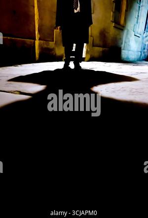 Eine Person in Anzug und Krawatte steht in einer schwach beleuchteten Gasse und wirft einen großen Schatten auf den Boden. Stockfoto