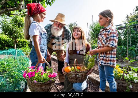 Bauernmutter hält einen Korb voll mit frisch gepflückten Früchten, während Töchter und Vater die pulsierende Ernte auf ihrer Bio-Farm bewundern und einen genießen Stockfoto