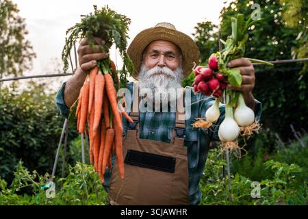 Lächelnder Seniorenbauer mit langem Bart und Strohhut, der stolz auf frisch geerntete Karotten, Radieschen und Zwiebeln in seinem lebhaften Obstgarten, Embod, zeigt Stockfoto