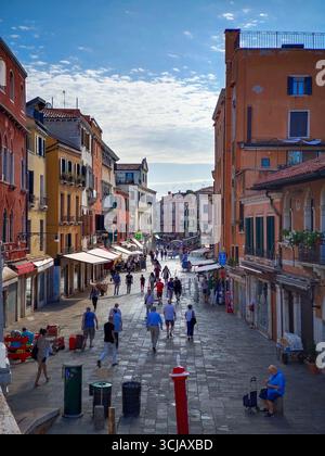 Eine Weitwinkelaufnahme einer überfüllten Straße in Venedig. Das Foto hebt die klassische Architektur und die kräftigen Farben der historischen Gebäude mit pe hervor Stockfoto