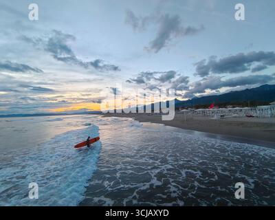 Eine atemberaubende Weitwinkelaufnahme des Strandes in Forte dei Marmi, Toskana. Das Foto zeigt einen Surfer mit einem roten Brett, der in Twilig im Meer spaziert Stockfoto