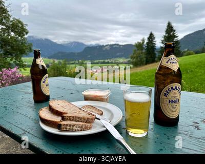 Zwei Flaschen Meckatzer Höllenbier und ein Glas Roggensauerteigbrot mit Frischkäse auf grünem Tisch mit den Alpen im Hintergrund Stockfoto